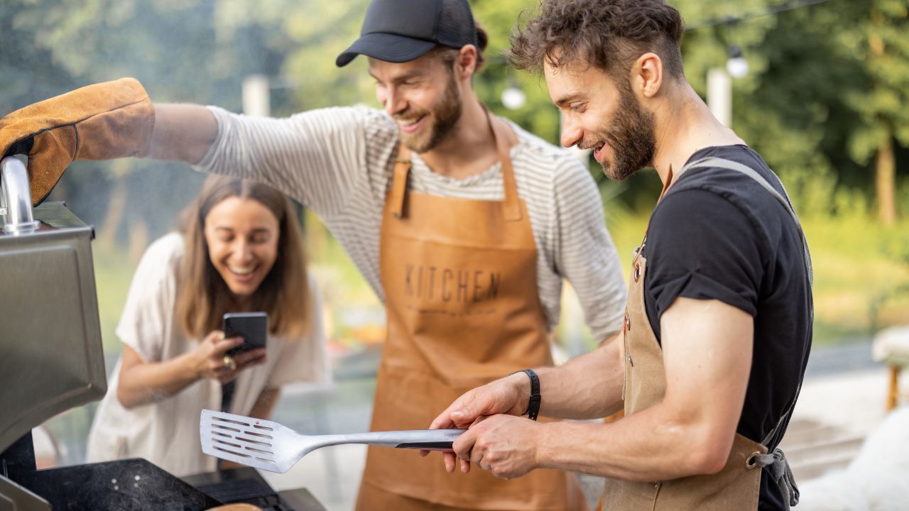 Zwei Männer grillen gemeinsam im Freien, während Freunde den Sommer genießen. Das Bild vermittelt gesellige Grillabende und entspannte Zeit mit Familie und Freunden.
