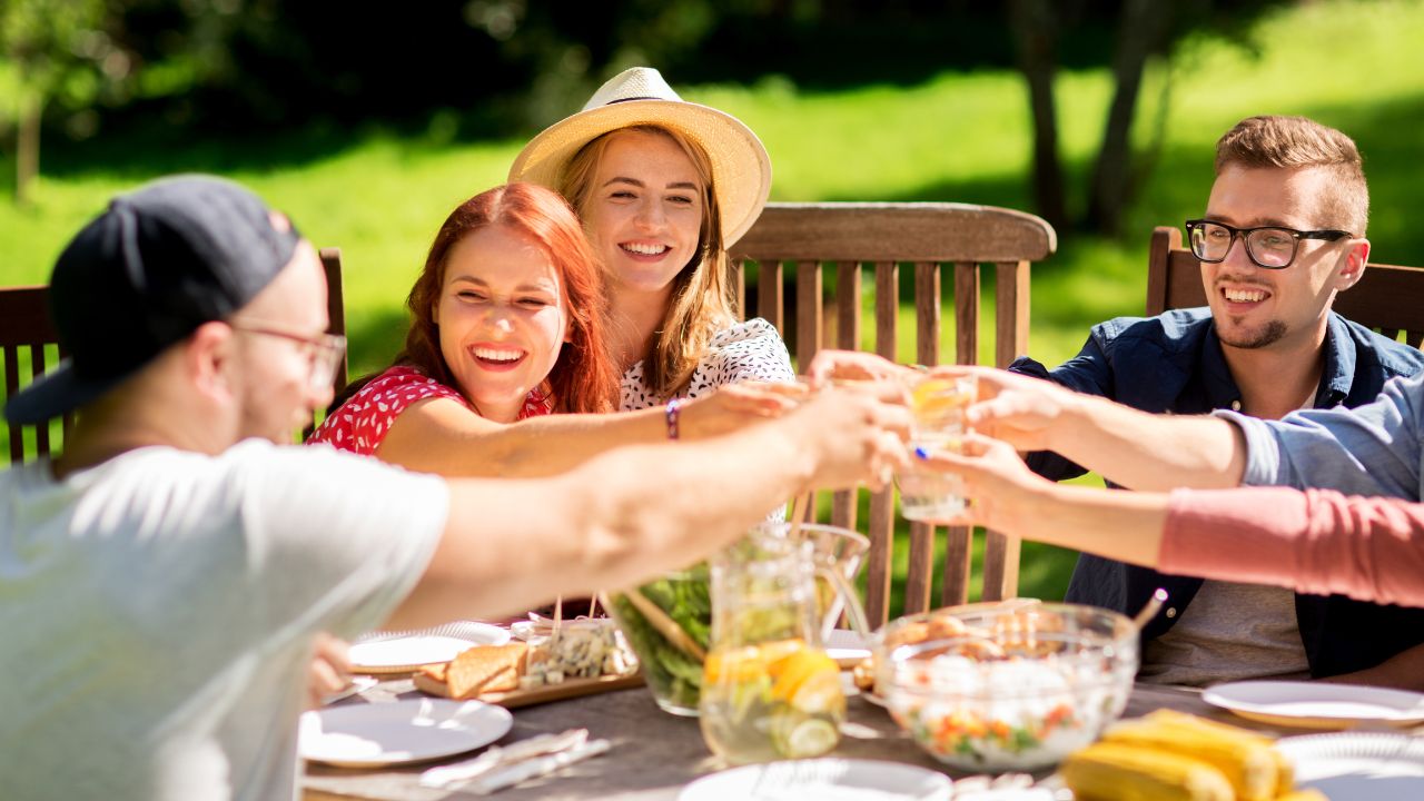 Des amis trinquent autour d'une table dressée dans un jardin ensoleillé. La table est garnie de plats, de boissons et d'ingrédients frais, dans une ambiance conviviale et estivale.