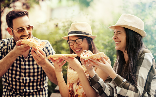 Freunde sitzen gemeinsam im Freien, essen und lachen miteinander. Das Bild zeigt eine entspannte sommerliche Atmosphäre bei einem gemeinsamen Essen mit Freunden.